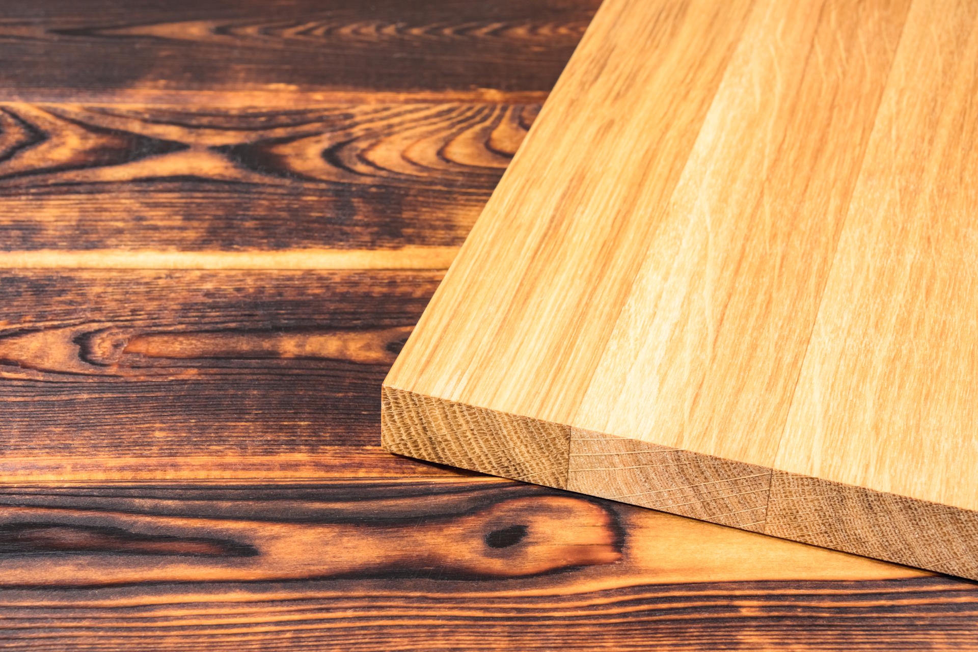 Plattenwerkstoff Die Plattenwerkstoffe von HolzPartner in Rathenow bieten höchste Qualität für Möbelbau, Innenausbau und kreative Projekte in Berlin und Brandenburg. Dank robuster Verarbeitung und natürlicher Holzoptik schaffen sie langlebige und ästhetische Lösungen. Bildbeschreibung: Helle Holzplatte mit klarer Maserung liegt auf einem dunklen, gebrannten Holzuntergrund mit markanter Struktur.