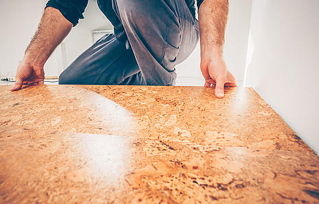 Korkboden Korkboden von HolzPartner in Rathenow – perfekte Lösung für ein nachhaltiges und behagliches Zuhause in Berlin und Brandenburg. Kork ist fußwarm, elastisch und sorgt für ein angenehmes Raumklima – ideal für Wohn- und Schlafbereiche. Bildbeschreibung: Handwerker verlegt Korkboden in einem Raum, Nahaufnahme der natürlichen Korkstruktur.