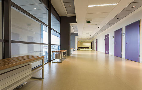 Linoleumboden von HolzPartner in Rathenow – die strapazierfähige und umweltfreundliche Bodenlösung für Berlin und Brandenburg. Linoleum ist pflegeleicht, langlebig und eignet sich perfekt für stark beanspruchte Räume wie Flure, Büros oder öffentliche Einrichtungen.  Bildbeschreibung: Moderner Flur mit großflächigem Linoleumboden in heller Farbe, mehreren Türen und Sitzgelegenheiten.