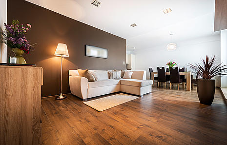 Massivholzdielen Massivholzdielen von HolzPartner bringen natürliche Wärme, Eleganz und Beständigkeit in Ihr Zuhause. Jede Diele besteht aus einem Stück Holz und sorgt für ein unvergleichliches Wohnambiente mit zeitloser Ausstrahlung. Robust, langlebig und mit individueller Maserung verleihen sie Wohnräumen in Berlin und Brandenburg ein Stück echte Natur. Bildbeschreibung: Moderner Wohnraum mit dunkelbraunen Massivholzdielen, heller Eckcouch, Essbereich und stilvoller Einrichtung.