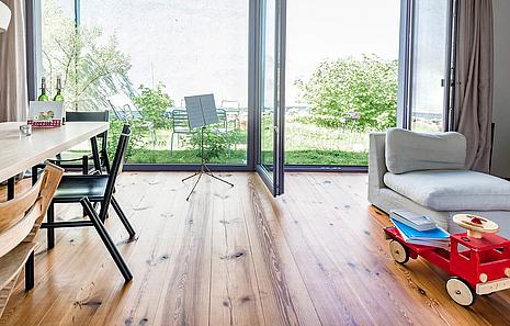 Massivholzdielen Massivholzdielen von HolzPartner vereinen Natürlichkeit und Beständigkeit – für ein gesundes Wohnklima und ein zeitlos schönes Zuhause. Jede Diele ist ein Unikat mit einzigartiger Maserung und bringt die Natur direkt in Ihre Wohnräume in Berlin und Brandenburg. Bildbeschreibung: Moderner Wohn- und Essbereich mit großen Glasfronten, hellen Massivholzdielen, Esszimmermöbeln und Kinderspielzeug im gemütlichen Ambiente.