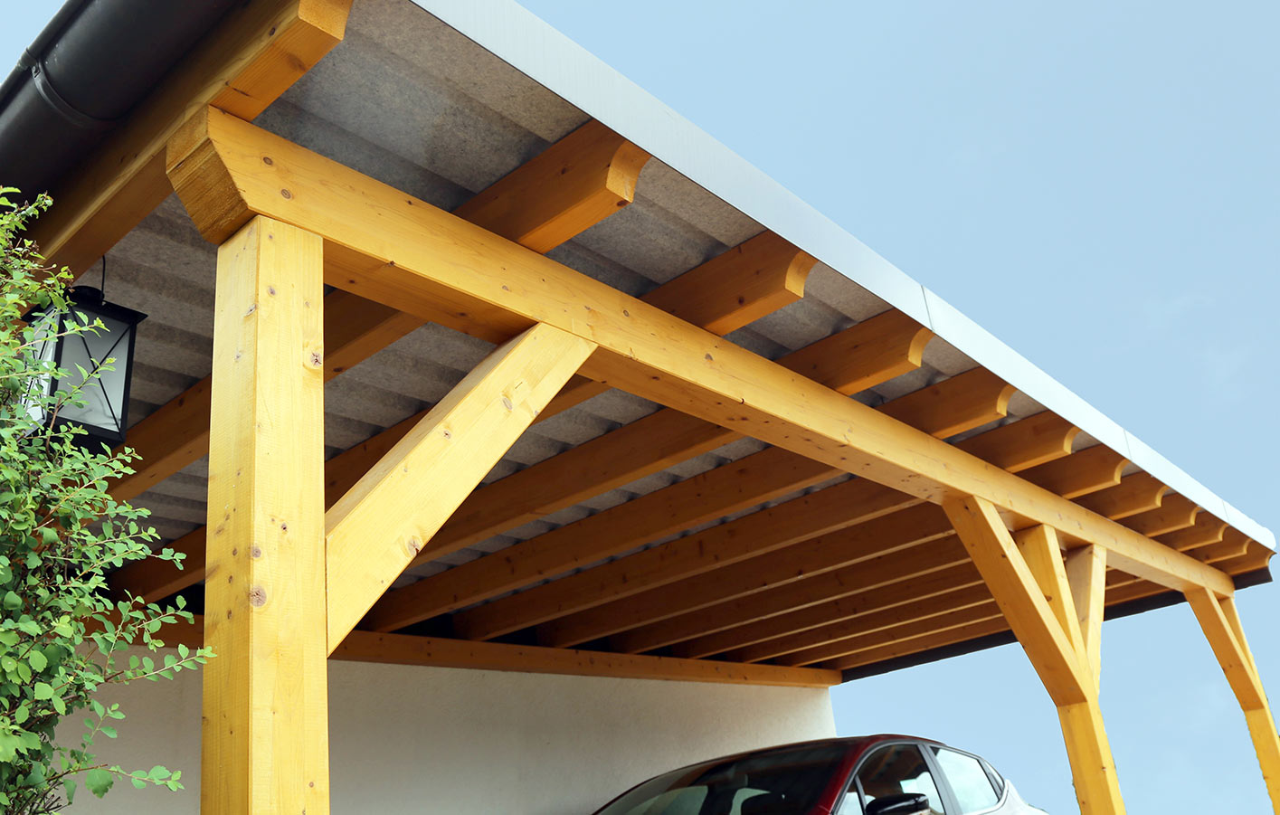 HolzPartner in Rathenow bietet stabile und langlebige Carports aus Holz für Berlin und Brandenburg – die ideale Lösung, um Ihr Auto vor Regen, Schnee und Sonne zu schützen. Wählen Sie aus verschiedenen Designs, die Funktionalität und ansprechende Optik verbinden.  Bildbeschreibung: Holz-Carport mit massivem Gestell und Flachdach, darunter ein geparktes Auto, seitlich eine Hauswand und Begrünung.