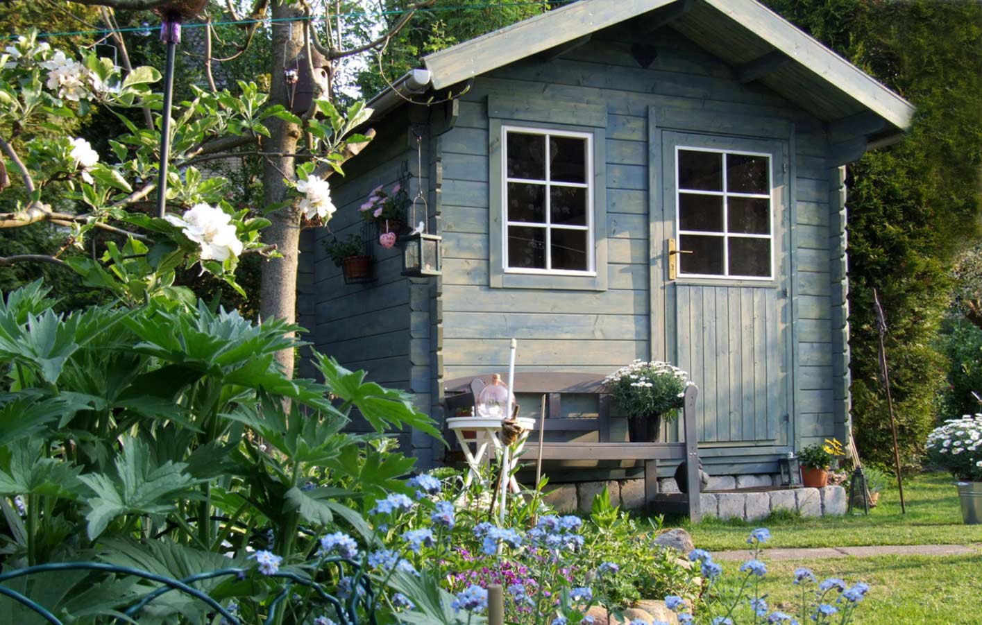HolzPartner in Rathenow bietet hochwertige Gartenhäuser für Berlin und Brandenburg – ideal als Stauraum, Rückzugsort oder dekoratives Highlight im Garten. Setzen Sie auf langlebiges Holz und vielseitige Designs für Ihren Außenbereich.  Bildbeschreibung: Kleines Gartenhaus aus Holz mit Satteldach, Fenster und Tür, davor eine Bank, Blumen und grüne Pflanzen im Garten.