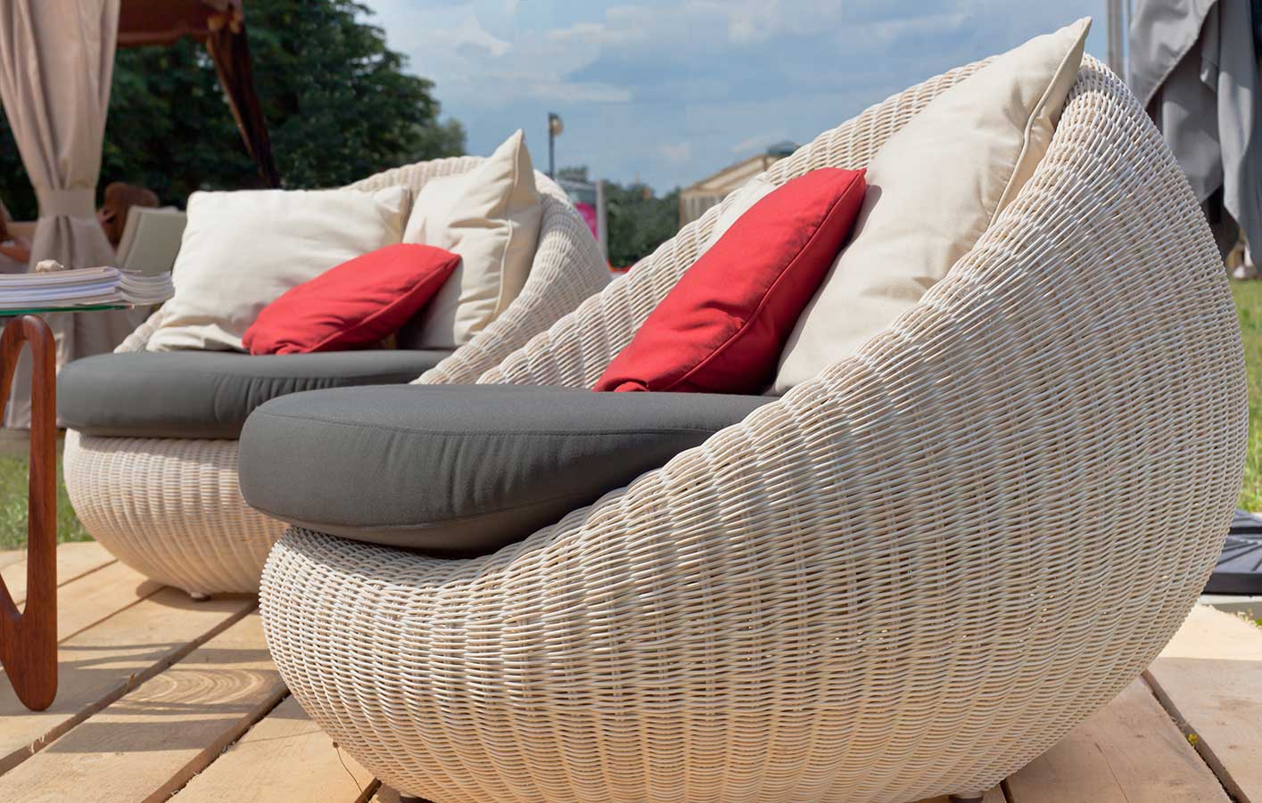 Mit stilvollen Lounge-Sesseln von HolzPartner in Rathenow gestalten Sie in Berlin und Brandenburg Ihre Terrasse oder den Garten zu einem Ort der Entspannung. Bequem, wetterfest und modern – ideal für gemütliche Stunden im Freien.  Bildbeschreibung: Zwei runde, geflochtene Lounge-Sessel in Weiß mit grauen Sitzpolstern und roten sowie beigen Kissen, aufgestellt auf einer Holzterrasse im Freien.