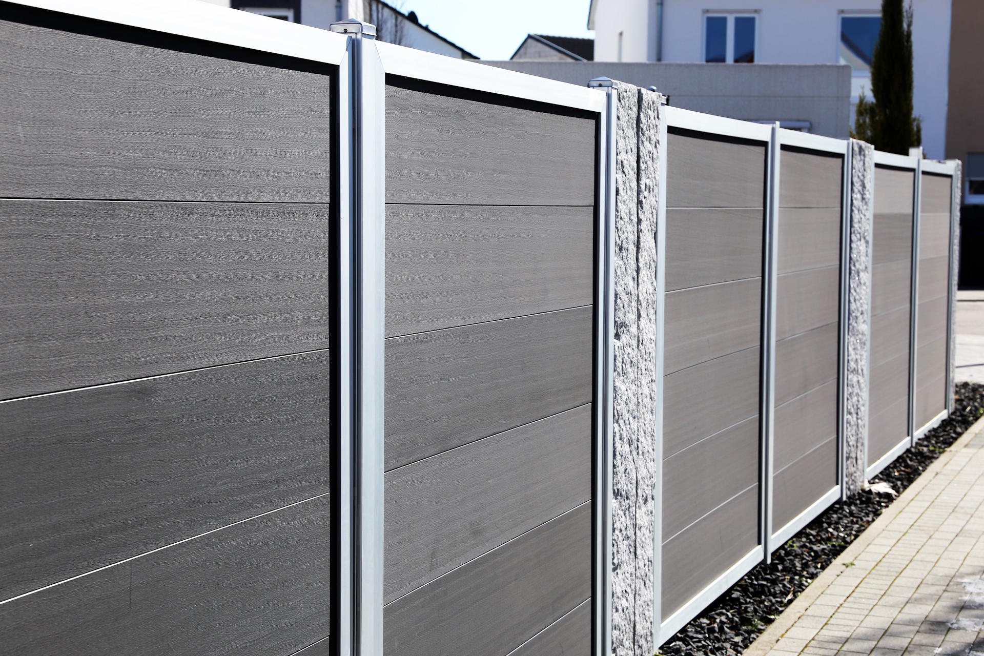 Ein Sichtschutzzaun aus WPC von HolzPartner in Rathenow bietet Haus- und Gartenbesitzern in Berlin und Brandenburg eine langlebige, pflegeleichte und moderne Lösung für Privatsphäre und Design. Wetterfest und stilvoll fügt er sich harmonisch in jede Umgebung ein.  Bildbeschreibung: Moderner Sichtschutzzaun aus grauen WPC-Elementen mit silberfarbenen Metallpfosten und seitlich integriertem Steinpaneel.