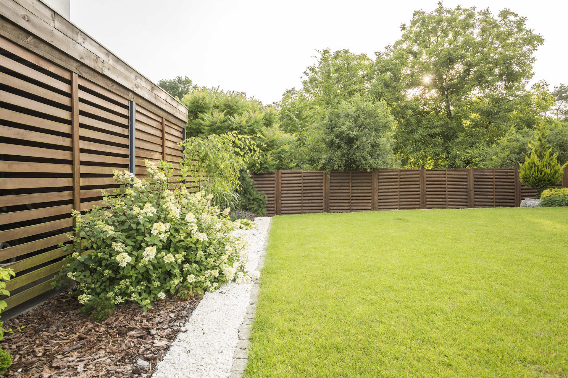Ein Sichtschutzzaun aus Holz von HolzPartner in Rathenow ist die natürliche Lösung für Haus und Garten in Berlin und Brandenburg. Er verbindet Privatsphäre mit warmer, zeitloser Optik und fügt sich harmonisch in jede Außenanlage ein.  Bildbeschreibung: Moderner Sichtschutzzaun aus horizontalen, dunkelbraun lasierten Holzbrettern mit hellen Pfosten, aufgestellt entlang eines Gehwegs und Gartens.