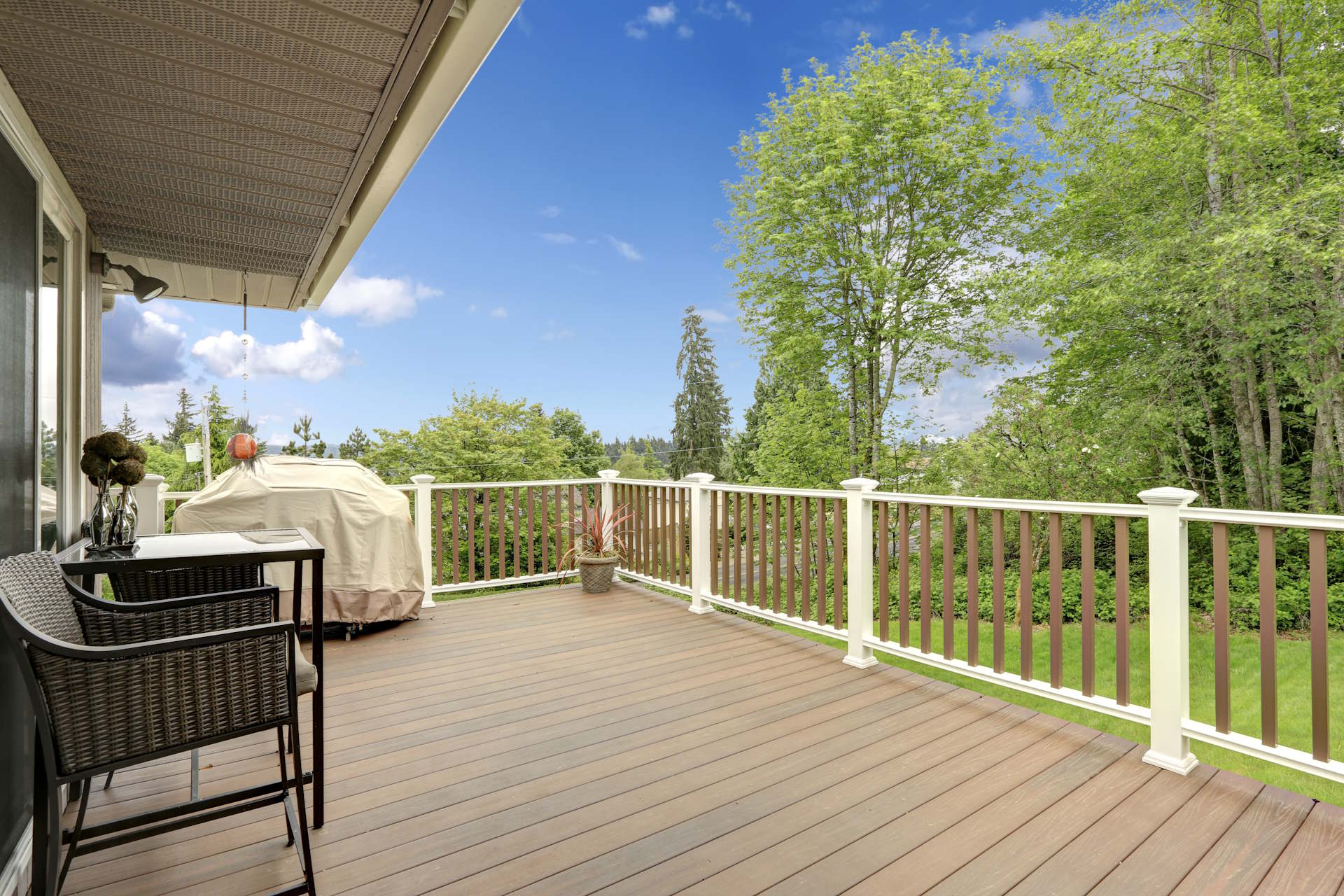 Terrassendielen aus Holz HolzPartner in Rathenow bietet Terrassendielen aus Holz für Berlin und Brandenburg – robust, wetterbeständig und in natürlicher Optik. Schaffen Sie mit hochwertigen Holzdielen eine einladende Terrasse für entspannte Stunden im Freien. Bildbeschreibung: Überdachte Terrasse mit braunen Holzdielen, weiß-braunem Geländer, Gartenmöbeln und Blick auf einen grünen Garten mit Bäumen.