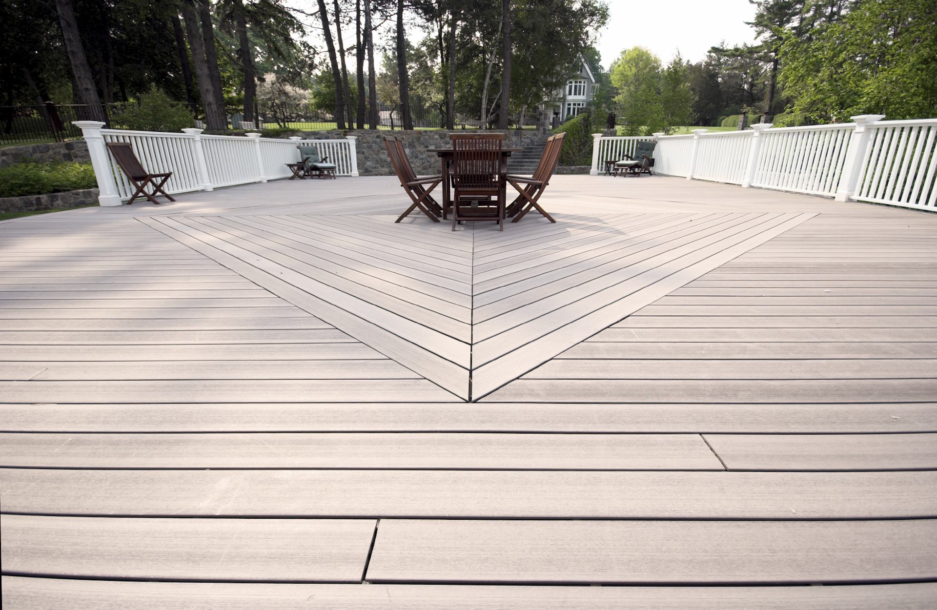 Terrassendielen aus WPC HolzPartner in Rathenow bietet Terrassendielen aus WPC für Berlin und Brandenburg – langlebig, pflegeleicht und ideal für stilvolle Außenbereiche. Genießen Sie die Kombination aus moderner Optik und hoher Widerstandsfähigkeit gegenüber Witterungseinflüssen. Bildbeschreibung: Große Terrasse mit grauen WPC-Dielen, weißen Geländern und einer Sitzgruppe aus Holzstühlen und -tisch in der Mitte.