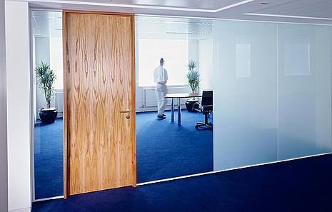 Innentüren Innentüren von HolzPartner in Rathenow bringen Stil und Funktionalität in Ihr Zuhause oder Büro in Berlin und Brandenburg. Sie sind in vielfältigen Designs und Oberflächen erhältlich, sodass sie perfekt zu jedem Wohn- oder Arbeitskonzept passen. Bildbeschreibung: Moderne Innentür aus Holz in einer hellen Büroumgebung mit Glaswänden, blauem Teppichboden und einem Mann im Hintergrund.