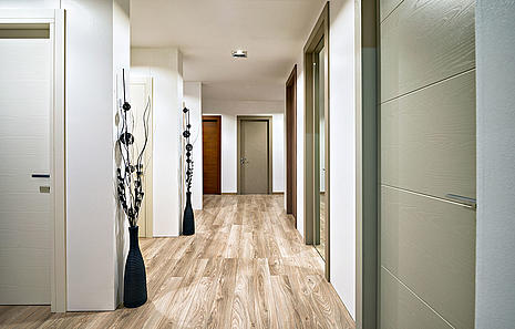 Innentüren In der Innentüren-Ausstellung von HolzPartner in Rathenow entdecken Kunden aus Berlin und Brandenburg eine große Auswahl an modernen Türdesigns. Unterschiedliche Oberflächen, Farben und Materialien bieten individuelle Gestaltungsmöglichkeiten für jeden Wohnstil. Bildbeschreibung: Moderner Flur mit verschiedenen Innentüren in hellen und dunklen Holztönen, dekorativen Vasen und Holzfußboden.