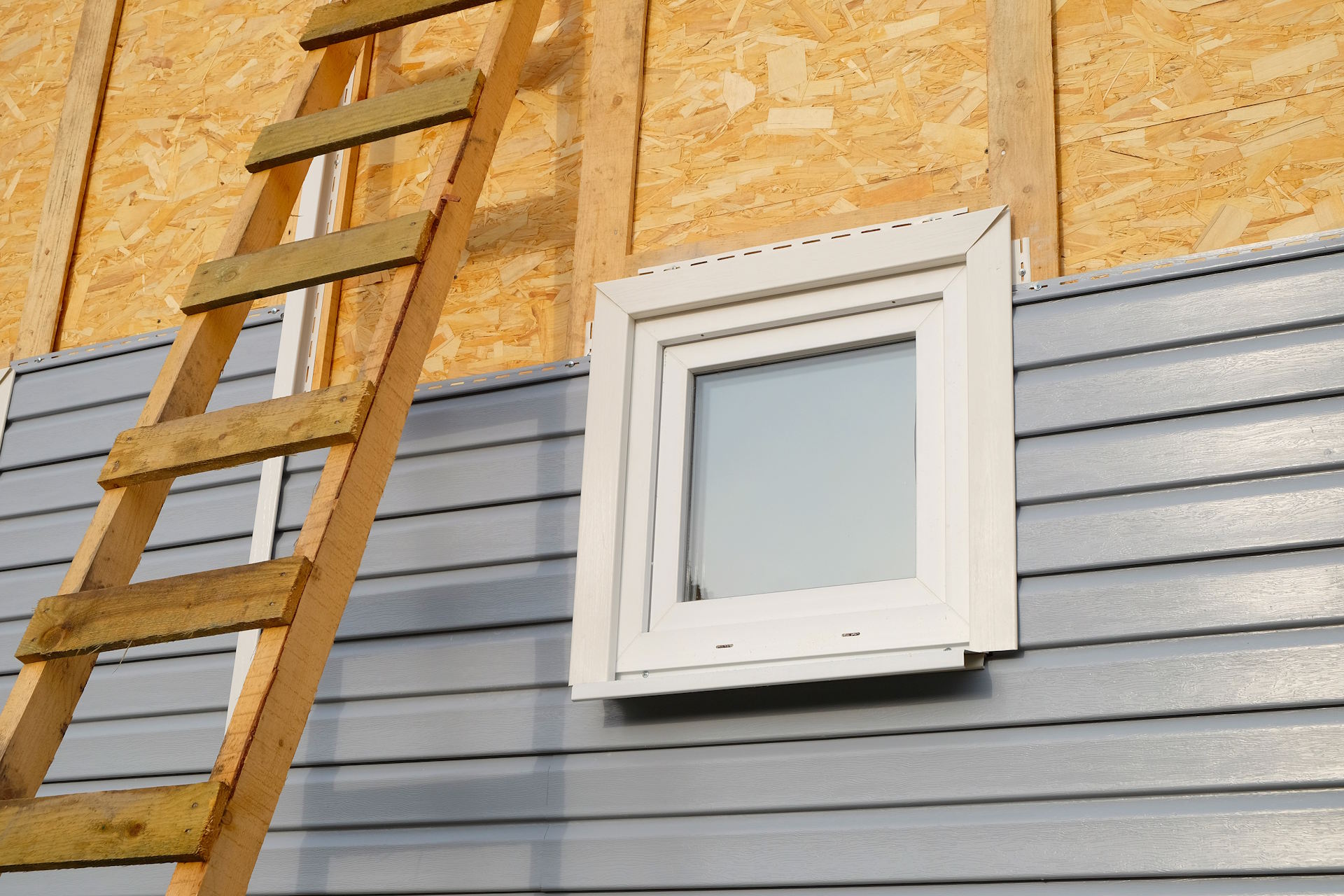 Holzfassade Mit einer Holzfassade von HolzPartner in Rathenow setzen Sie in Berlin und Brandenburg auf natürliche Ästhetik und energieeffizienten Schutz für Ihr Zuhause. Die Fassade ist robust, nachhaltig und lässt sich individuell gestalten – für ein langlebiges und stilvolles Ergebnis. Bildbeschreibung: Hauswand im Bau mit grauer Holzfassade in horizontaler Optik, einem weißen Fensterrahmen und einer angelehnten Holzleiter.