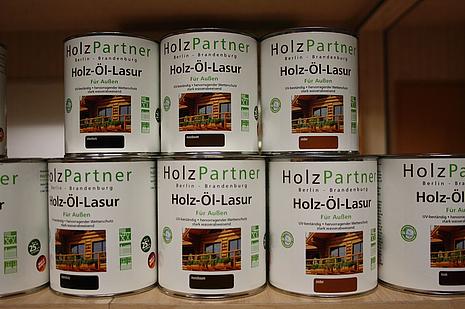 Holzschutz Die Holzschutz Farben und Lasuren von HolzPartner in Rathenow bieten langlebigen Schutz und eine natürliche Optik für Außenholz in Berlin und Brandenburg. Sie schützen zuverlässig vor Witterungseinflüssen und verleihen Ihrem Holz eine hochwertige, pflegende Oberfläche. Bildbeschreibung: Mehrere weiß etikettierte Dosen Holz-Öl-Lasur mit Aufdruck „HolzPartner“ stehen im Regal, abgebildet mit Hausmotiv und Farbmustern in Braun- und Holztönen.