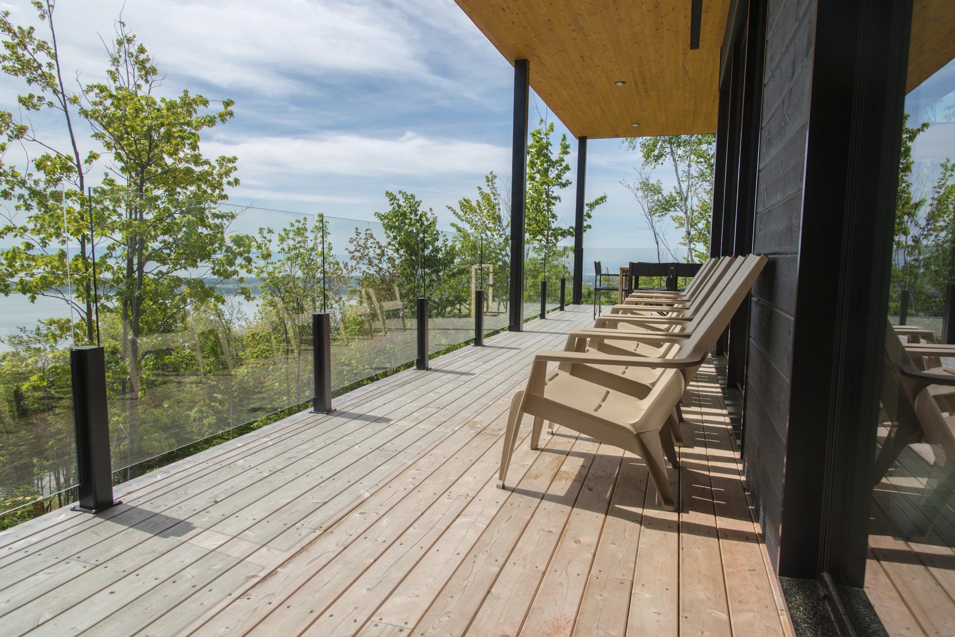 Terrassendielen aus Holz HolzPartner in Rathenow bietet langlebige Terrassendielen aus Holz für Berlin und Brandenburg – wetterbeständig, hochwertig und in stilvollen Designs. Gestalten Sie Ihre Terrasse mit natürlicher Ausstrahlung und genießen Sie Qualität, die begeistert. Bildbeschreibung: Holzterrasse mit Glasgeländer, mehreren Kunststoffstühlen und Ausblick auf Bäume sowie Landschaft im Hintergrund.
