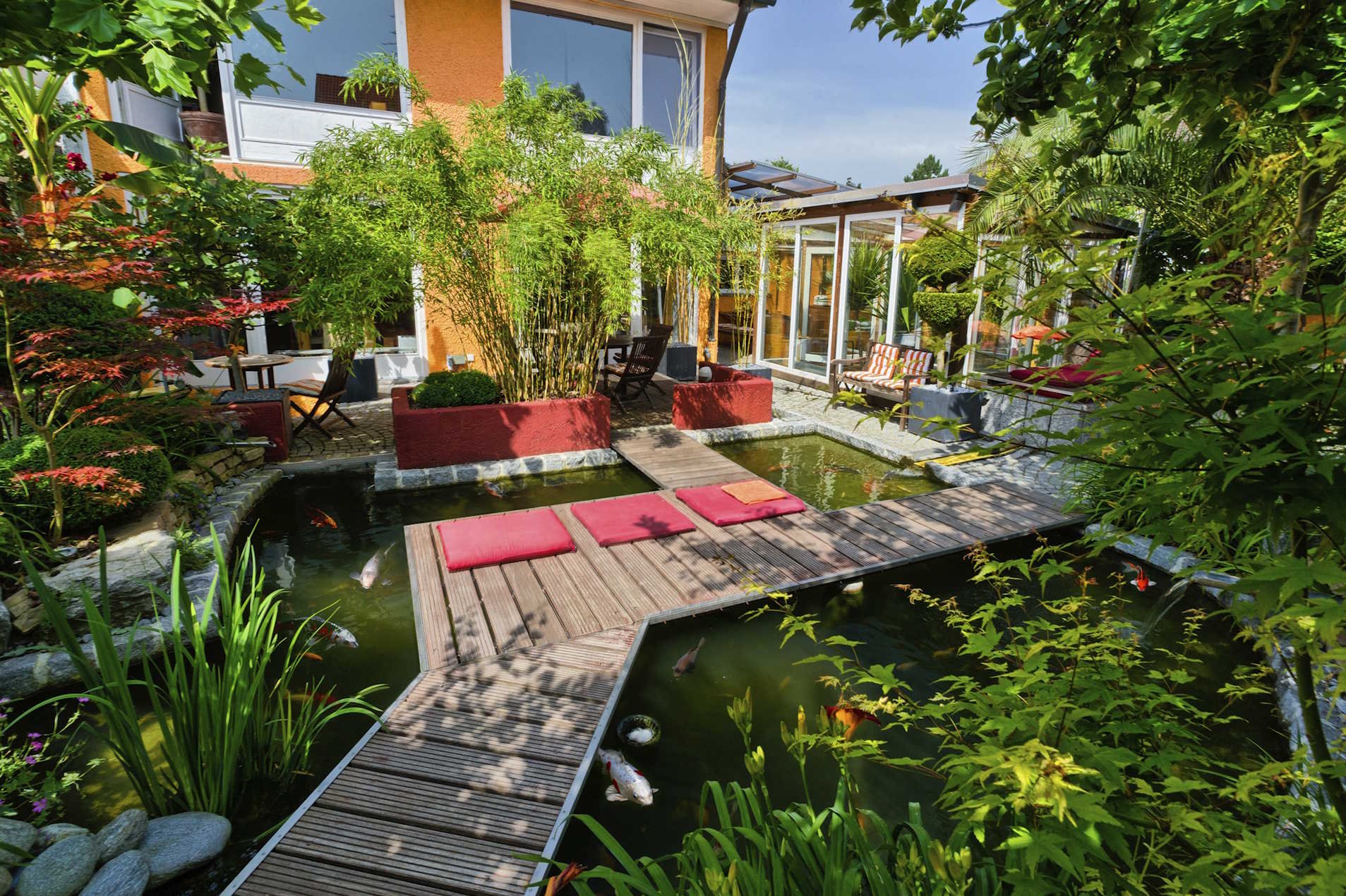 Terrassendielen aus Holz HolzPartner in Rathenow bietet hochwertige Terrassendielen aus Holz für Berlin und Brandenburg – langlebig, wetterfest und ideal zur Gestaltung von Garten, Balkon oder Poolbereich. Genießen Sie natürliche Ästhetik und strapazierfähige Qualität für Ihren Außenbereich. Bildbeschreibung: Garten mit Holzterrasse und Teich, darauf rote Sitzkissen sowie umliegende Pflanzen und Gartenmöbel an einem Wohnhaus.