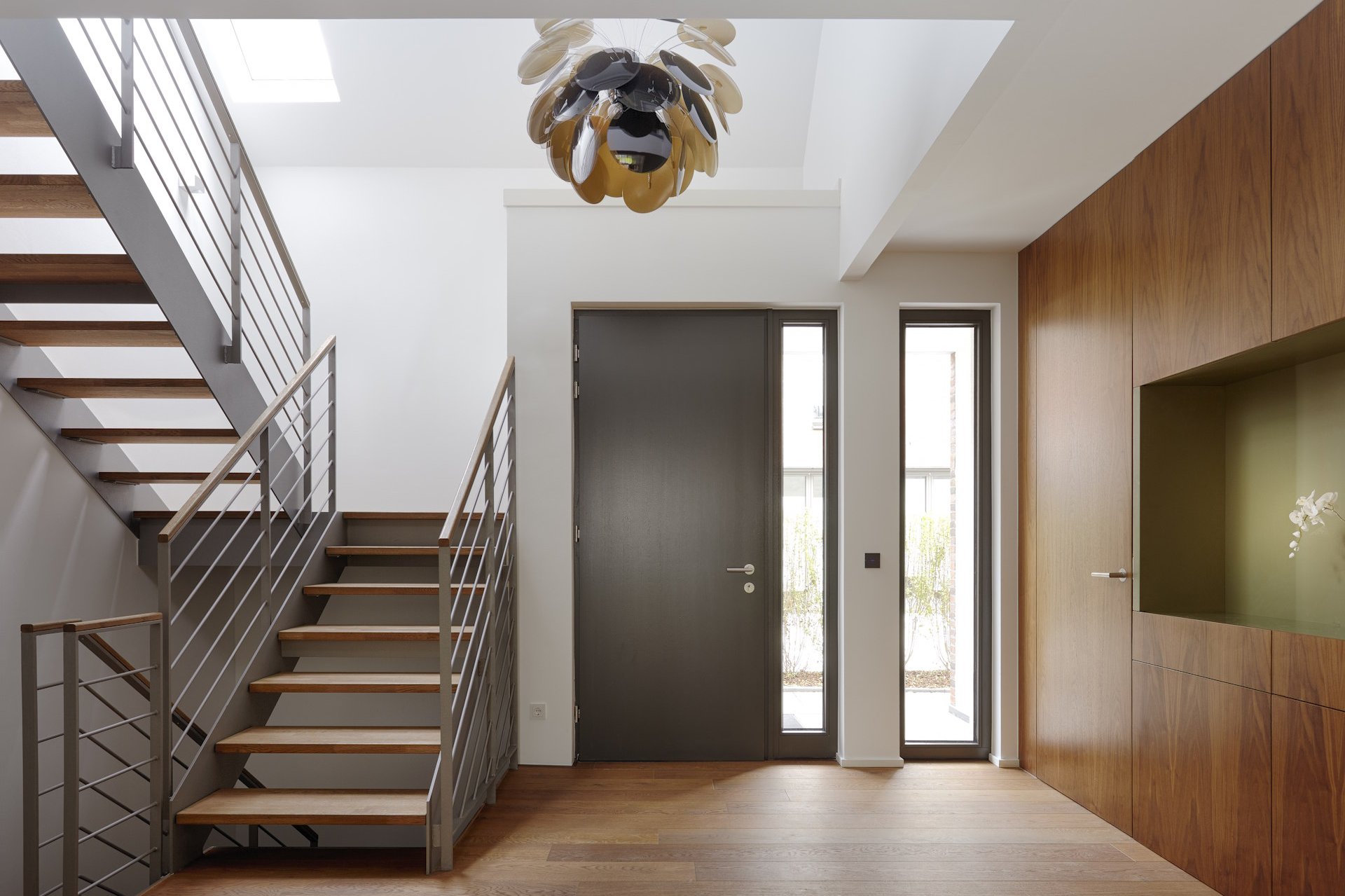 Haustüren In der Haustüren Ausstellung von HolzPartner in Rathenow entdecken Sie eine große Auswahl an modernen und klassischen Türen für Berlin und Brandenburg. Ob in elegantem Holzdesign, mit Glas-Elementen oder in zeitloser Farbe – hier finden Sie die perfekte Tür, die Sicherheit, Funktionalität und stilvolles Design vereint. Bildbeschreibung: Moderner Eingangsbereich mit einer dunklen Haustür, seitlichen Glaselementen, Holzverkleidung an der Wand und offener Treppe mit Holzstufen und Metallgeländer.