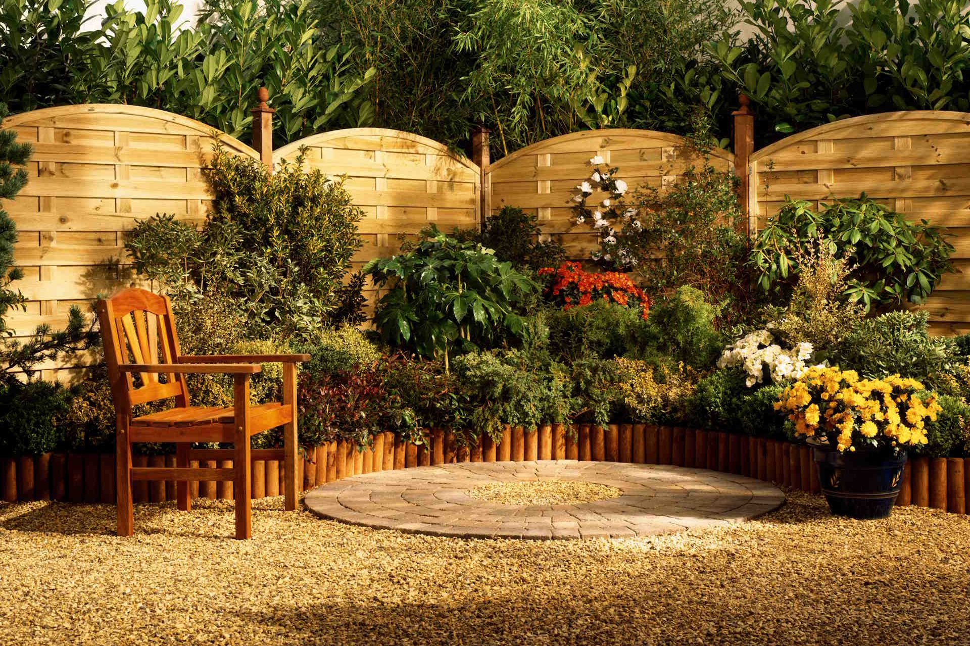 Sichtschutzelemente von HolzPartner in Rathenow schaffen in Berlin und Brandenburg eine geschützte Wohlfühlatmosphäre im Garten. Die Holzelemente bieten Privatsphäre, wirken zugleich natürlich und setzen dekorative Akzente.  Bildbeschreibung: Garten mit geschwungenen Sichtschutzelementen aus hellem Holz, davor bunte Blumenbeete, Sträucher und ein einzelner Holzstuhl auf Kiesfläche.