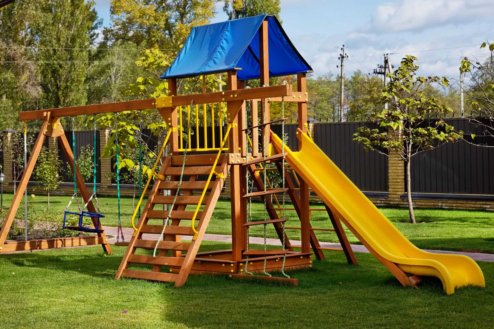 Kinderspielgeräte für den Garten HolzPartner in Rathenow bietet für Familien in Berlin und Brandenburg hochwertige Kinderspielgeräte aus robustem Holz. Mit Schaukeln, Rutschen und Klettertürmen verwandeln Sie Ihren Garten in einen sicheren Spielplatz, der Kinder zu Bewegung und Abenteuern einlädt. Bildbeschreibung: Holzspielgerät im Garten mit gelber Rutsche, blauem Dach, Kletterelementen und Schaukeln auf einer gepflegten Rasenfläche.