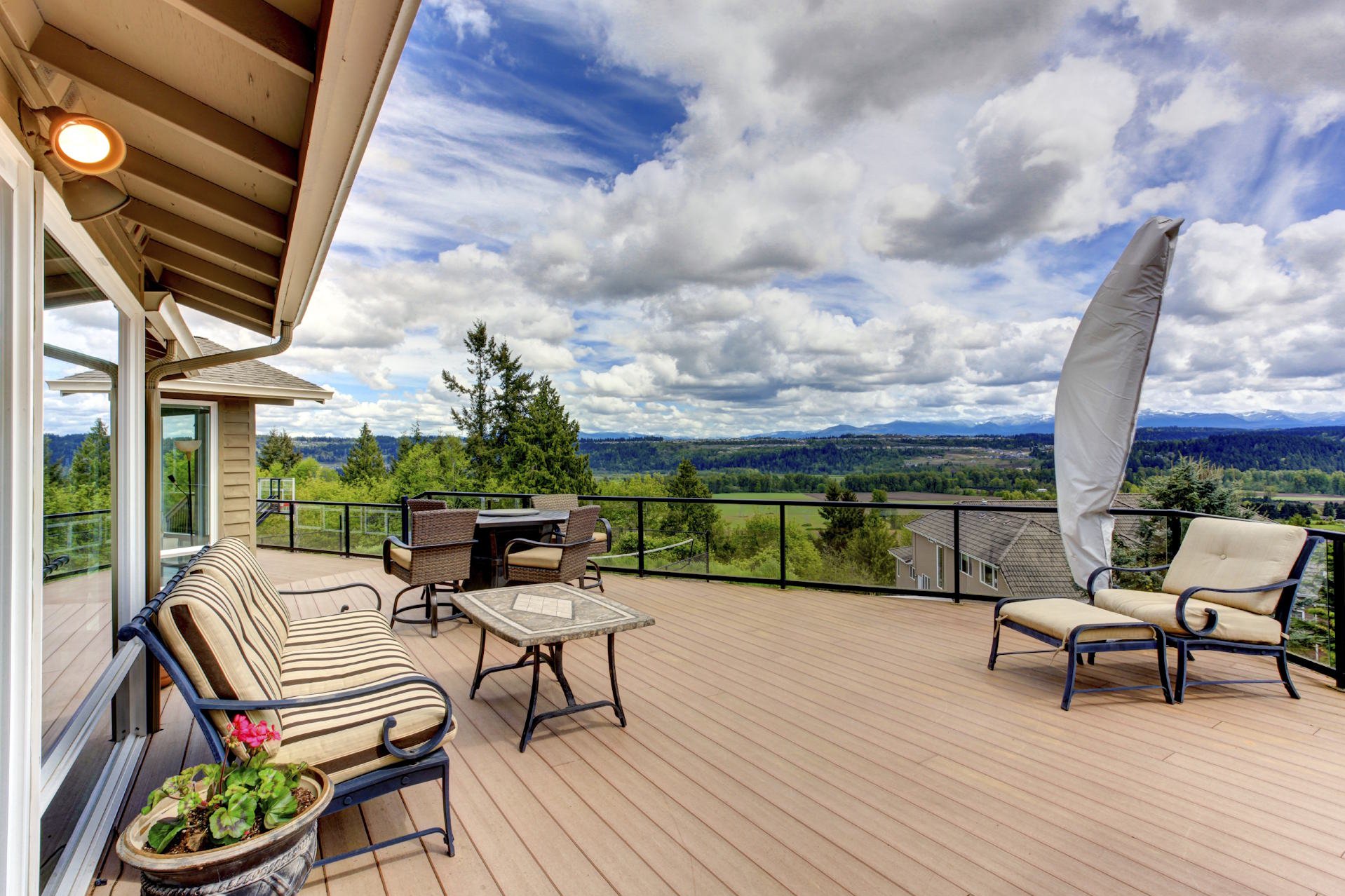 Terrassendielen aus WPC HolzPartner in Rathenow bietet pflegeleichte Terrassendielen aus WPC für Berlin und Brandenburg – wetterfest, langlebig und in moderner Holzoptik. Gestalten Sie Ihre Terrasse komfortabel und genießen Sie stilvolles Wohnen im Außenbereich. Bildbeschreibung: Große Terrasse mit braunen WPC-Dielen, Sitzmöbeln mit Polstern, Tisch und weitem Ausblick über Landschaft, Bäume und Himmel.